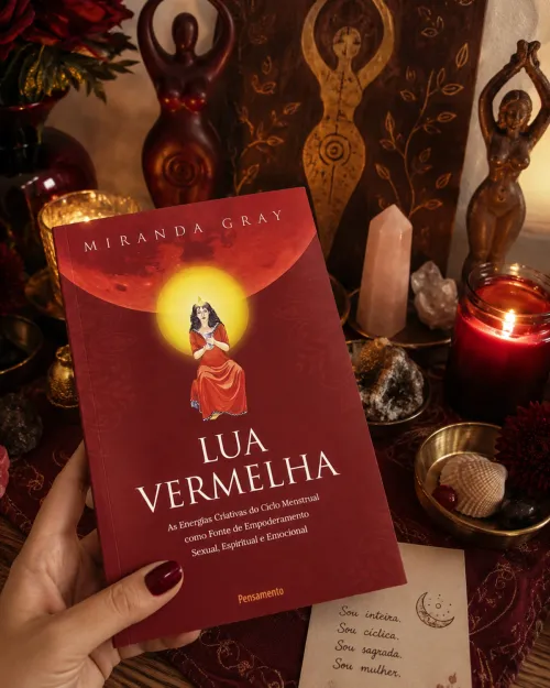 Mãos segurando o livro Lua Vermelha diante de um altar com velas e estatuetas